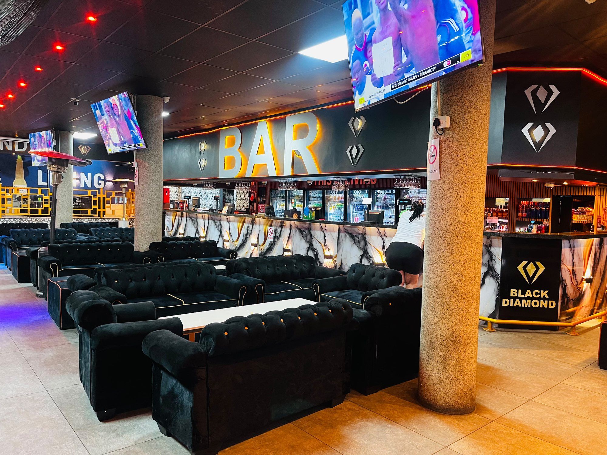 The long bar seen across the seating: an illuminated BAR sign in gold above stocked fridges, a marble-patterned bar front lit from below, and a concrete pillar in the foreground.