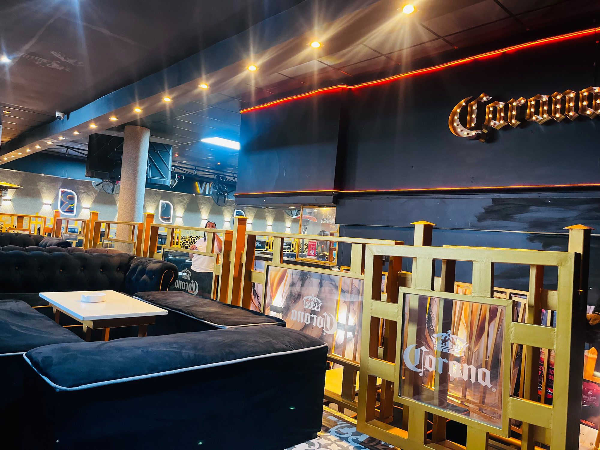 A raised seating area behind a gold screen, with a lit Corona marquee sign in bulb lettering on the dark wall above and a low white table in the foreground.