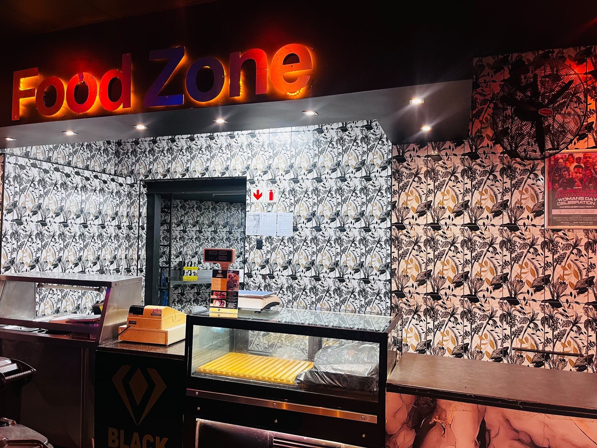 The Food Zone counter at Black Diamond: an illuminated FOOD ZONE sign in red and blue above a stainless serving hatch, with a glass display cabinet and patterned monochrome wallpaper behind.