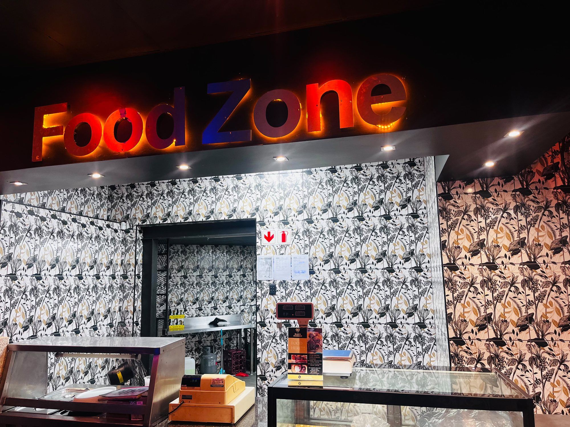 The Food Zone counter: an illuminated sign in red and blue lettering above a serving hatch, patterned monochrome wallpaper, a glass display cabinet and a till.