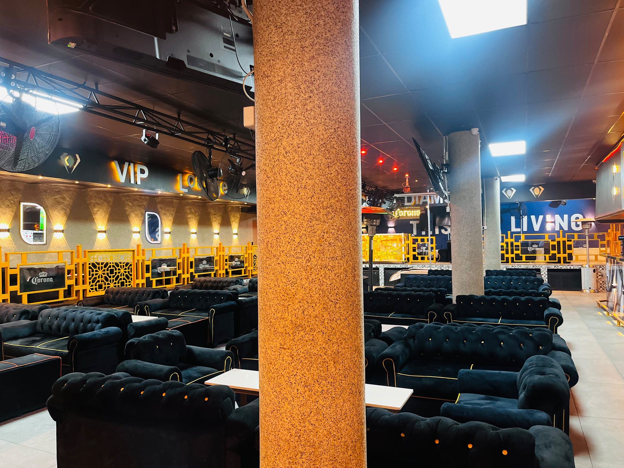 Black buttoned velvet armchairs and low white tables arranged around a stone-clad pillar, with a gold balustrade and Corona-branded panels behind.