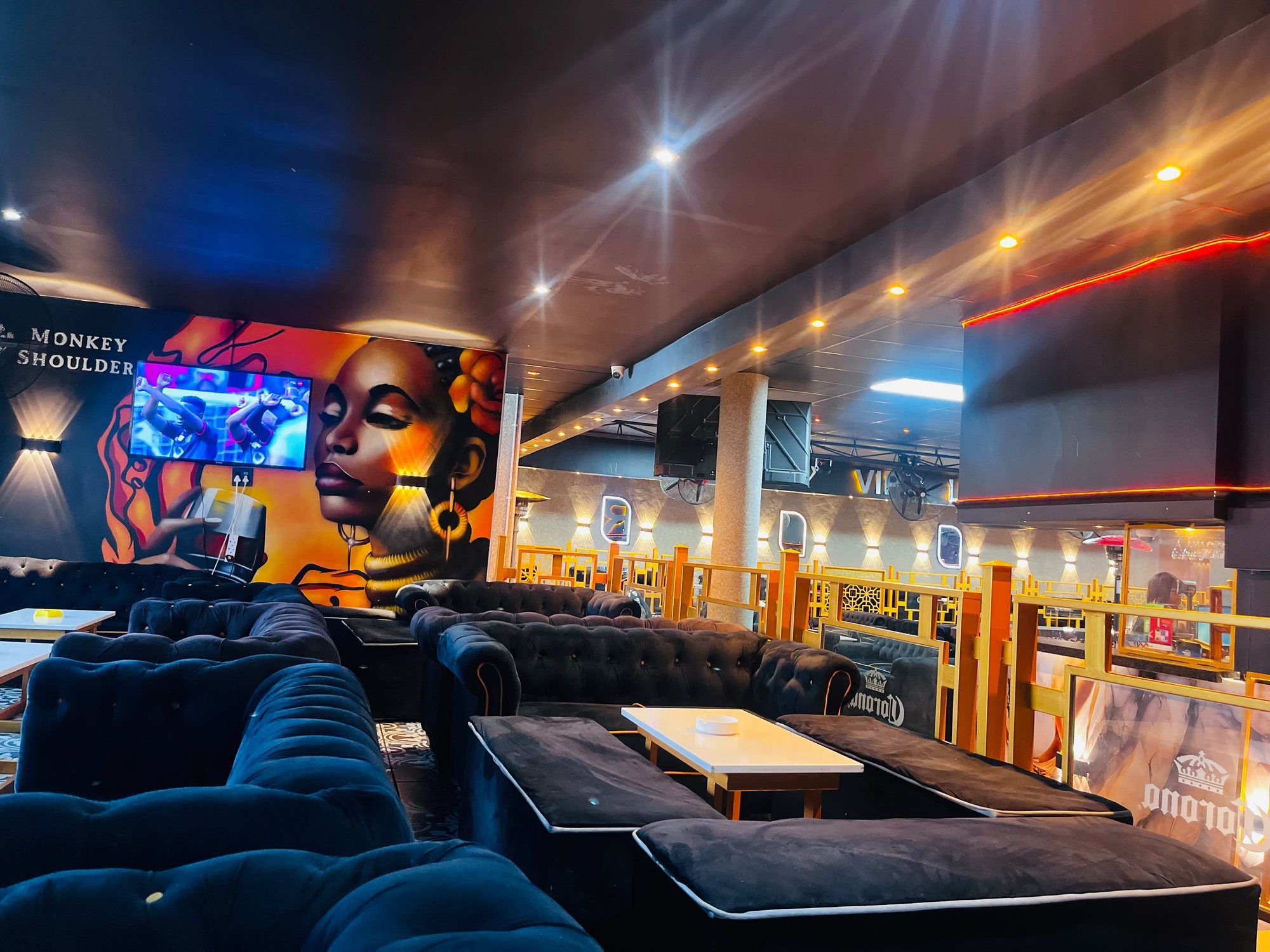 A painted mural of a woman in profile wearing a gold headwrap and hoop earrings, beside a wall-mounted screen and the Monkey Shoulder wordmark, with velvet seating below.